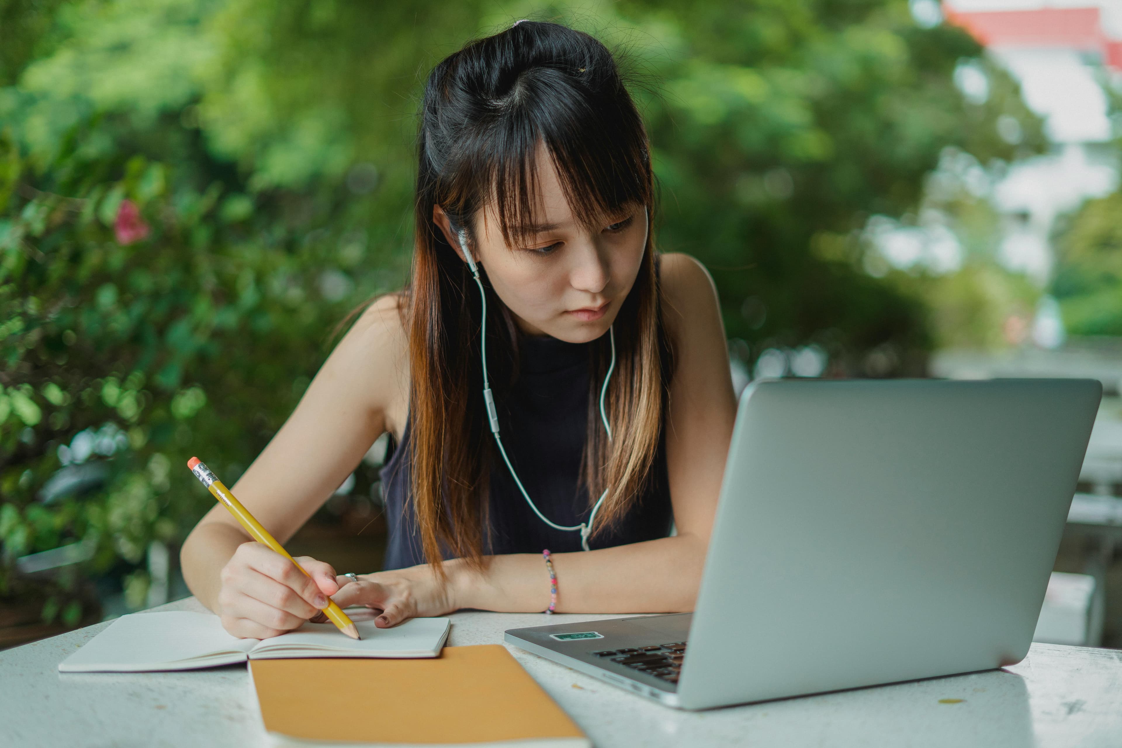 A young woman wearing earphones, focused on her laptop while taking notes in a notebook, symbolizing the dedication required for studying marketing data analytics in a serene outdoor environment.