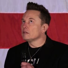 Elon Musk stares upwards in front of a US flag.