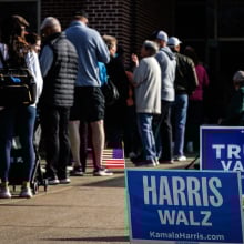 Harris and Trump yard signs are staked next to a line of voters entering a polling place.