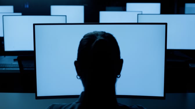 The back of a woman's head, as she stares at several glowing blue screens.